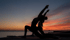 Zwei silhouettierte Personen machen Yoga in einer Ausfallschritt-Pose bei Sonnenuntergang am Meer.