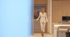A woman in a white suit opens a door to a modern kitchen with light wood cabinets.