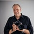 Der lächelnde Businessfotograf Oliver Reetz steht mit ei einer professionelle DSLR-Kamera mit großem Objektiv vor grauem Hintergrund.