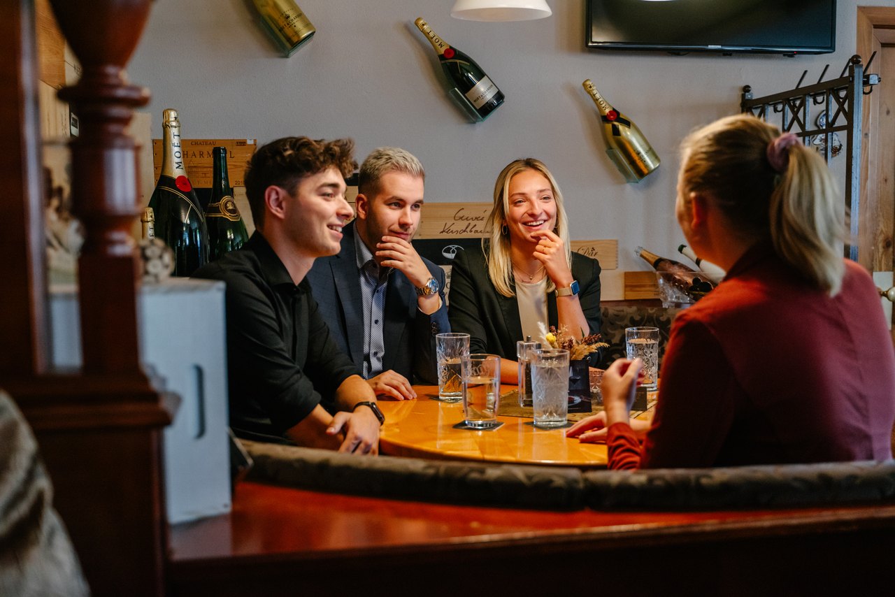Vier junge Leute sitzen an einem Holztisch in einer Bar mit Champagnerflaschen an der Wand.