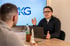 Ein Mann spricht mit einem anderen Mann an einem Tisch, dahinter ein Bildschirm mit "KKG Consulting" Logo.
