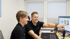 Zwei lächelnde Kollegen in schwarzen Poloshirts arbeiten am Schreibtisch und zeigen auf einen Computerbildschirm.