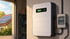Weiße Solarbatterie an einer Wand in einer Garage, daneben ein Sicherungskasten und ein Haus mit Solardach.