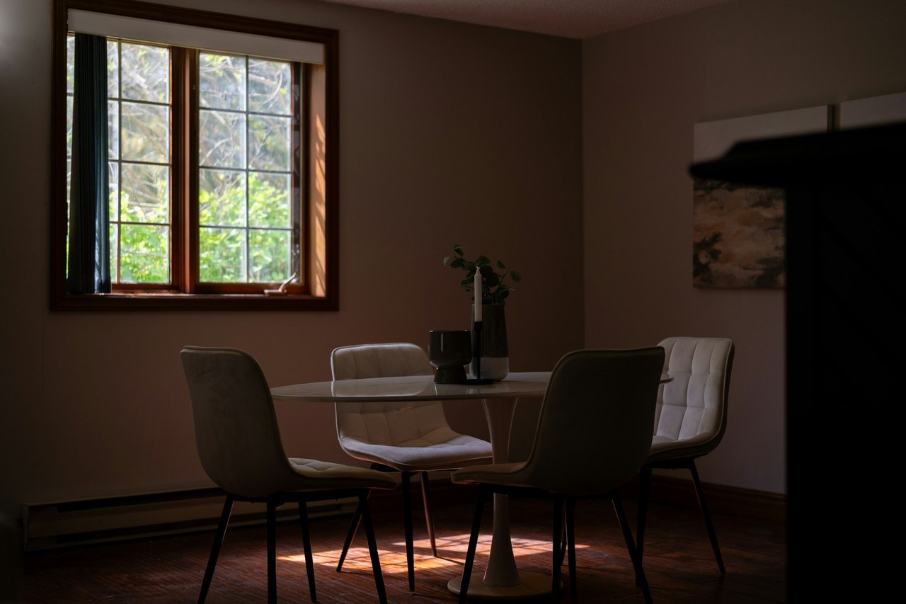 Ein dämmriges Esszimmer mit rundem Tisch und vier Stühlen, durch das Fenster fällt Licht auf den Boden.