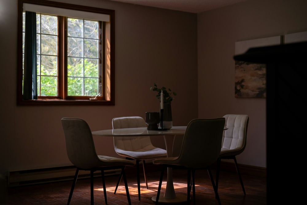 Ein dämmriges Esszimmer mit rundem Tisch und vier Stühlen, durch das Fenster fällt Licht auf den Boden.