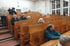 Menschen sitzen in einem leeren Hörsaal mit Holzstühlen und Tischen, einige schauen nach vorne.