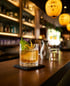Signature Cocktail mit Yuzu-Zeste und Shiso-Blatt auf dem Bartresen des Hikari, dahinter die leuchtenden Lampions und Flaschenregale
