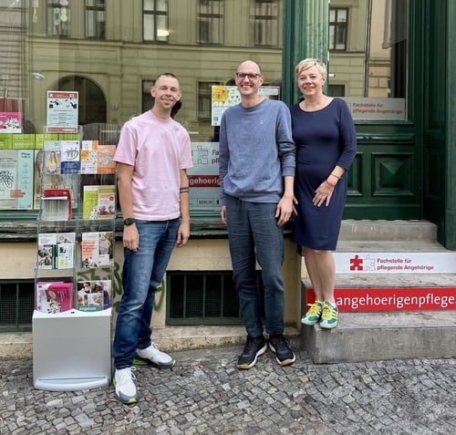 Three people stand in front of a building with the inscription “Fachstelle für Angehörigenpflege”.