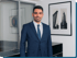 Ein Mann im blauen Anzug und Krawatte lächelt, vor einem Büro mit gerahmten Gebäudefotos.