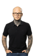 Kahlköpfiger Mann mit Brille und Tattoos in schwarzem Poloshirt und Jeans.