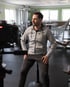 Kunde im grauen Hoodie und dunkler Hose sitzt auf einem Hocker in einem Fitnessstudio, seitlich zur Kamera für ein Imagefilm-Interview.