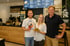 Drei Gastronomen vor moderner Café-Theke mit digitaler Menütafel im Hintergrund, halten Kaffee-to-go und Sandwich – Teamportrait für Gastronomie-Websites