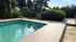Außenpool mit türkisfarbenem Wasser und einer hellen Poolumrandung aus WPC-Dielen in einem Garten mit Bäumen und Hecken.