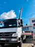 Weißer Mercedes-Benz LKW mit Arbeitsbühne und ausgefahrenem Ausleger vor einem Gebäude unter blauem Himmel.