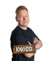 Porträt von Knossi mit verschränkten Armen vor schwarzem Hintergrund und einem Holzschild mit der Aufschrift KNOSSI.