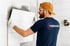 Ein Mann in orangefarbener Mütze und blauem "THERMAHEAT"-Shirt verlegt weiße Fliesen an einer Wand.