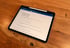 Ein schwarzes Tablet zeigt eine Liste mit dem Titel "Benötigte Unterlagen" auf einem Holztisch.
