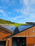Zwei Holzdächer mit Solarpaneelen überragen eine grüne Wiese mit Waldrand unter blauem Himmel.