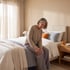 Senior woman sits on the edge of a bed holding her knee with a pained expression.