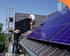 Zwei Arbeiter installieren Solarmodule auf einem dunklen Ziegeldach.