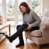 A woman in a gray sweater sits in an armchair, holding her thigh with a pained expression.