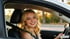 Eine lächelnde junge Frau mit blonden Haaren sitzt angeschnallt am Steuer eines Autos bei Sonnenuntergang.