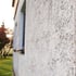 Textured white stucco wall of a house with a blurred window, green grass, and red foliage in the background.