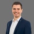 Tom Hanker, professionelles Business-Porträt eines lächelnden Mannes im dunkelblauen Anzug mit weißem Hemd vor neutralem grauen Hintergrund.