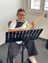 Smiling girl with glasses sitting on a chair, holding a light green electric guitar, next to a music stand.
