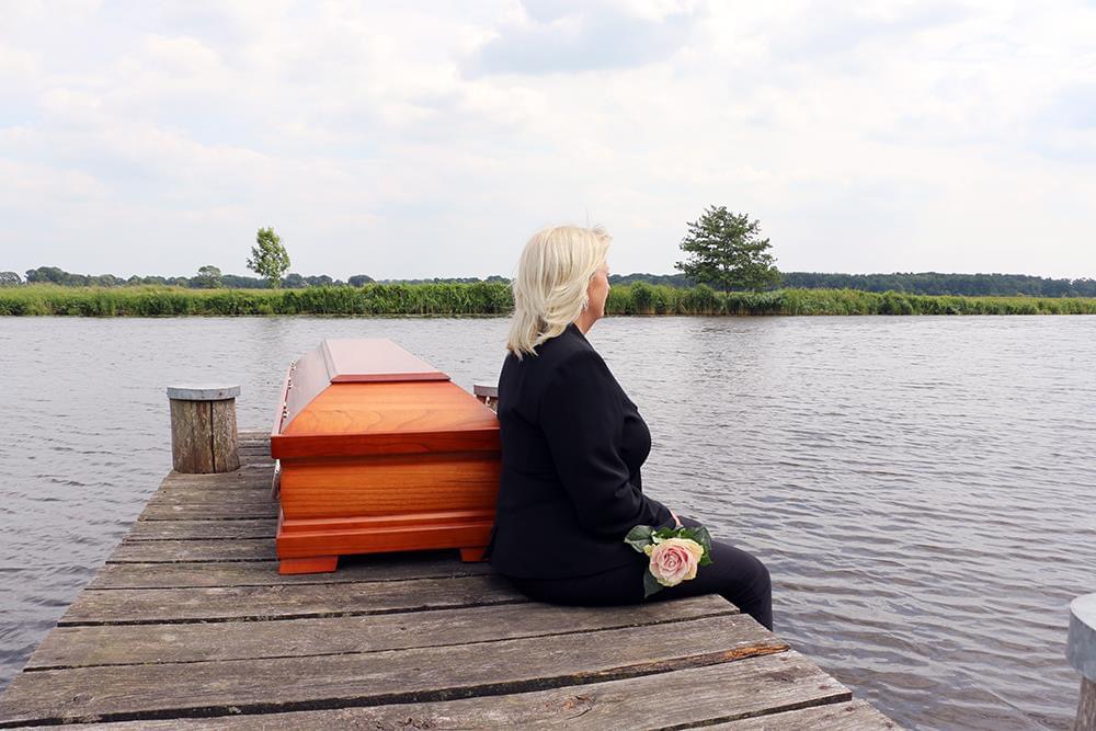 Trauernde Frau sitzt am Steg neben einem Sarg am ruhigen Wasser – Abschied bei einer Seebestattung