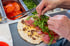 Hände legen Rucola auf ein offenes Fladenbrot mit Schinken, Käse und Olivenpaste. Im Hintergrund geschnittene Tomaten.