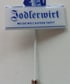 Ein weißer Anstecker mit der Aufschrift „Jodlerwirt WO DIE WELT BAYERN TRIFFT“ in blauer Schrift.