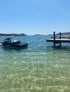 Ein schwarzer Jet-Ski schwimmt auf klarem, grünlichem Wasser neben einem Holzsteg. Im Hintergrund sind Hügel und Inseln.