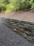 Natursteinmauer aus dunklen Grauwacke-Blöcken vor einem Hang mit frischer Erde und grüner Bepflanzung.