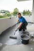 Ein Handwerker in blauem T-Shirt und Schutzbrille versiegelt einen Balkonboden mit grauer Farbe.