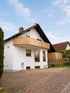Weißes Einfamilienhaus mit Holz-Balkon, gepflasterter Einfahrt und einer Wallbox-Ladestation an der Außenwand.