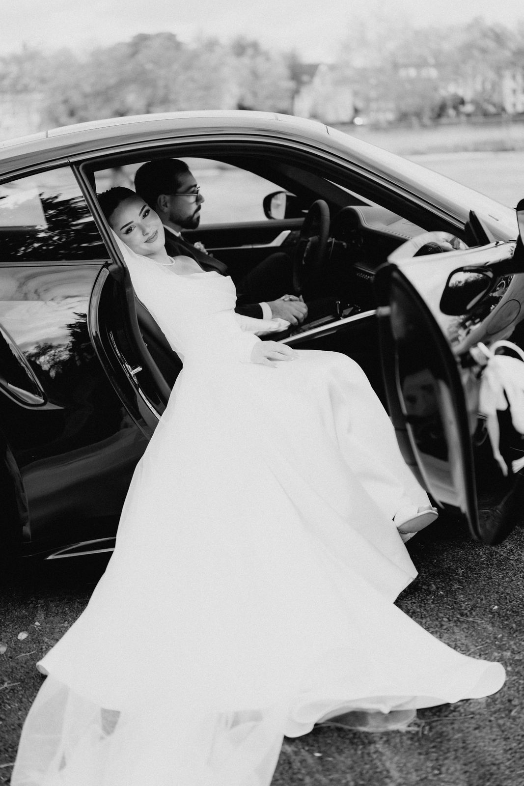 Braut sitzt im geöffneten Hochzeitsauto vor Schloss Benrath in Düsseldorf, das Kleid fällt aus der Tür, Schwarzweißaufnahme.