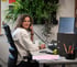Eine Frau mit lockigem Haar telefoniert am Schreibtisch in einem Büro mit vielen Zimmerpflanzen.