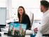 Zwei Kollegen lächeln und sprechen an Schreibtischen in einem modernen Büro mit Computern.