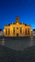 Die Ludwigskirche in Saarbrücken bei Nacht, beleuchtet und vor einem tiefblauen Himmel. Der gepflasterte Vorplatz ist zu sehen.