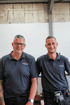 Andreas und Norman Singer in dunkelblauen Poloshirts mit Firmenlogo stehen nebeneinander in einer Werkstatt.