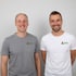 Zwei lächelnde Männer in T-Shirts mit dem Weder-Logo stehen vor einer hellgrauen Wand.