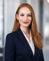 Lachende Frau mit roten Haaren, rotem Lippenstift, dunklem Blazer und weißer Bluse vor verschwommenem Hintergrund.