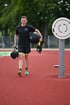 Mann in Sportkleidung geht auf rotem Sportplatz, trägt einen schwarzen Medizinball und eine Sporttasche.