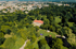 Luftaufnahme eines roten Gebäudes umgeben von grünen Bäumen und Rasenflächen mit einer Stadt im Hintergrund.