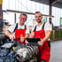 Zwei Mechaniker in roten Overalls und weißen T-Shirts lächeln, während sie an einer Maschine in einer Werkstatt arbeiten.