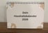 Beiger Tischkalender mit Spiralbindung und Text "Dein Haushaltskalender 2026", dekoriert mit braunen Blättern.