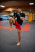 Ein Mann in roten Shorts und schwarzen Boxhandschuhen posiert in einer Muay-Thai-Kampfhaltung mit erhobenem Knie in einem Fitnessstudio.