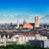Blick auf die Münchner Frauenkirche und Stadtlandschaft mit schneebedeckten Alpen im Hintergrund unter blauem Himmel.