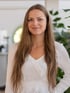 Junge Frau mit langen braunen Haaren und weißem Oberteil lächelt in die Kamera.
